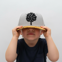 Ein kleiner Junge sitzt vor einem hellgrauen Studiohintergrund und zieht sich mit beiden Händen eine graue Kappe mit einem Schirm in Holzoptik und einem schwarzen Baum-Logo über die Augen, sodass nur sein Mund sichtbar ist.