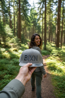 Aus der Ich-Perspektive hält eine Hand eine graue Kappe mit dem Schriftzug "BIXN" in die Kamera. Im unscharfen Hintergrund geht eine Frau auf einem Waldweg und blickt über die Schulter zurück, wobei sie spielerisch die Zunge herausstreckt.