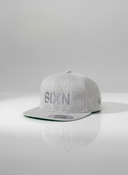 Ein professionelles Produktfoto einer grauen Kappe mit flachem Schirm. Auf der Vorderseite befindet sich der erhabene Schriftzug "BIXN" in einem Ton-in-Ton-Stich. Die Kappe steht auf einer spiegelnden Oberfläche vor einem hellen Studiohintergrund, was eine deutliche Reflexion erzeugt.