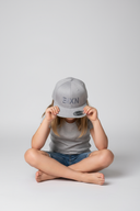 Ein barfüßiges Mädchen sitzt im Schneidersitz vor einem hellgrauen Studiohintergrund und hält sich mit beiden Händen eine graue Kappe vor das Gesicht. Die Kappe hat einen Ton-in-Ton-Schriftzug "BIXN".