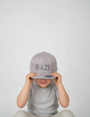Ein kleiner Junge sitzt vor einem hellgrauen Studiohintergrund und zieht sich mit beiden Händen eine graue Kappe über die Augen. Die Kappe hat einen Ton-in-Ton-Schriftzug "BAZI".