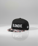Ein professionelles Produktfoto einer schwarzen Kappe mit flachem Schirm. Der Schirm hat ein buntes Blumenmuster und auf der Vorderseite steht der weiße Schriftzug "BLONDIE". Die Kappe steht auf einer spiegelnden Oberfläche vor einem hellen Studiohintergrund, was eine deutliche Reflexion erzeugt.