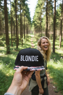 Aus der Ich-Perspektive hält eine Hand eine schwarze Kappe mit einem bunten Blumenschirm und dem weißen Schriftzug "BLONDIE" in die Kamera. Im unscharfen Hintergrund steht eine lachende Frau mit langen blonden Haaren auf einem sonnendurchfluteten Waldweg.