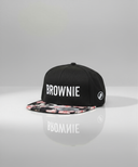 Ein professionelles Produktfoto einer schwarzen Kappe mit flachem Schirm. Der Schirm hat ein buntes Blumenmuster und auf der Vorderseite steht der weiße Schriftzug "BROWNIE". Die Kappe steht auf einer spiegelnden Oberfläche vor einem hellen Studiohintergrund, was eine deutliche Reflexion erzeugt.