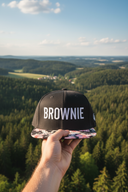 Aus der Ich-Perspektive hält eine Hand eine schwarze Kappe mit Blumenschirm und dem "BROWNIE"-Schriftzug in den Vordergrund. Im Hintergrund erstreckt sich eine weite, malerische Landschaft aus dichten Wäldern und sanften Hügeln.
