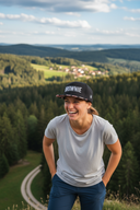 Eine fröhlich lachende Frau steht mit den Händen in den Hosentaschen auf einem Hügel. Sie trägt eine schwarze Kappe mit Blumenschirm und der Aufschrift "BROWNIE". Im Hintergrund erstreckt sich eine weite Landschaft aus bewaldeten Hügeln und einem kleinen Dorf.