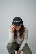 Eine lachende Frau mit langen, braunen Locken sitzt vor einem hellgrauen Studiohintergrund. Sie beugt sich nach vorne und zieht sich spielerisch eine schwarze Kappe mit dem weißen Schriftzug "BROWNIE" über die Augen.