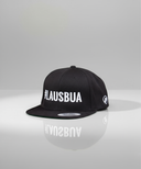 Ein professionelles Produktfoto einer schwarzen Kappe mit flachem Schirm. Auf der Vorderseite befindet sich der weiße, gestickte Schriftzug "#LAUSBUA". Die Kappe steht auf einer spiegelnden Oberfläche vor einem hellen Studiohintergrund, was eine deutliche Reflexion erzeugt.