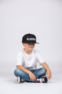 Ein kleiner Junge sitzt mit einem sanften Lächeln im Schneidersitz vor einem hellgrauen Studiohintergrund. Er trägt ein weißes T-Shirt, Jeans und eine schwarze Kappe mit dem weißen Schriftzug "#LAUSBUA".