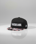 Ein professionelles Produktfoto einer schwarzen Kappe mit flachem Schirm. Der Schirm hat ein buntes Blumenmuster und auf der Vorderseite steht der weiße Schriftzug "#LAUSDEANDL". Die Kappe steht auf einer spiegelnden Oberfläche vor einem hellen Studiohintergrund, was eine deutliche Reflexion erzeugt.