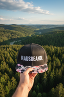 Aus der Ich-Perspektive hält eine Hand eine schwarze Kappe mit Blumenschirm und dem "#LAUSDEANDL"-Schriftzug in den Vordergrund. Im Hintergrund erstreckt sich eine weite, malerische Landschaft aus dichten Wäldern und einem Fluss im Tal, beleuchtet von der Abendsonne.