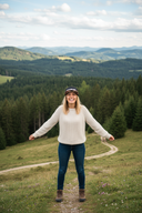 Eine fröhliche Frau in einem Strickpullover steht mit ausgebreiteten Armen auf einem Hügel. Sie trägt eine nach hinten gedrehte Kappe mit dem Schriftzug "#LAUSDEANDL" und blickt lachend in die Kamera. Im Hintergrund erstreckt sich eine weite Landschaft aus bewaldeten Hügeln.
