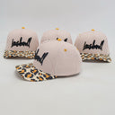 Vier identische Kinder-Kappen, die aus einer seitlichen Perspektive fotografiert wurden. Jede Kappe zeigt die Kombination aus einer gestreiften Krone und einem Schirm im Leopardenmuster vor einem weißen Studiohintergrund.