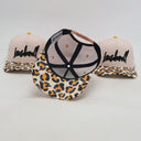 Ein Produktfoto von drei Kinder-Kappen mit "Lausdeandl"-Schriftzug, das verschiedene Details zeigt. Während zwei Kappen im Hintergrund die gestreifte Vorderseite präsentieren, ist die Kappe in der Mitte umgedreht, um die Unterseite des Schirms mit Leopardenmuster und den Lederriemen-Verschluss zu zeigen.