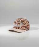 Ein professionelles Produktfoto einer Kinder-Kappe. Die Kappe hat ein Leopardenmuster, einen rosa-weiß gestreiften Schirm und den weißen, kursiven Schriftzug "Lausdeandl" auf der Vorderseite. Sie steht auf einer spiegelnden Oberfläche vor einem hellen Studiohintergrund, was eine deutliche Reflexion erzeugt.