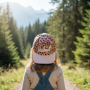 Ein Mädchen, von hinten fotografiert, steht auf einem Wanderweg in einem sonnigen Nadelwald. Es trägt eine Kappe mit Leopardenmuster und gestreiftem Schirm und blickt auf einen Berggipfel in der Ferne.