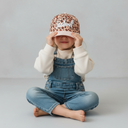 Ein barfüßiges Mädchen in einer Jeans-Latzhose sitzt im Schneidersitz vor einem hellgrauen Studiohintergrund. Es hält sich mit beiden Händen eine Kappe mit Leopardenmuster und gestreiftem Schirm vor das Gesicht und lächelt dabei.