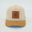 Ein professionelles Produktfoto einer Kappe in der Vorderansicht vor einem weißen Hintergrund. Die Kappe hat eine beige Krone aus Cordstoff und einen braunen Wildlederschirm. Im Zentrum befindet sich ein quadratischer Lederaufnäher mit einem geprägten Bergmotiv.