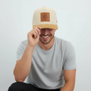 Ein Mann mit einem breiten Lächeln sitzt vor einem hellgrauen Studiohintergrund und zieht sich mit einer Hand spielerisch seine Kappe ins Gesicht. Die Kappe ist beige mit einem braunen Wildlederschirm und einem Lederaufnäher mit Bergmotiv.