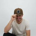 Ein Mann mit einem breiten Lächeln hockt vor einem hellgrauen Studiohintergrund und berührt den Schirm seiner olivgrünen Kappe. Die Kappe hat einen runden, schwarz-weißen "VOGEL-WUID"-Aufnäher auf der Vorderseite.