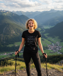 Eine Frau mit lockigen blonden Haaren lacht ausgelassen auf einem Berggipfel, während sie sich auf zwei Wanderstöcke stützt. Sie trägt ein schwarzes T-Shirt mit der Aufschrift "DA BERG RUAFT." vor einer atemberaubenden Kulisse aus einem grünen Tal und hohen Bergen.