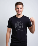 Ein junger Mann mit einem breiten, sympathischen Lächeln blickt direkt in die Kamera und trägt ein schwarzes T-Shirt mit dem weißen Aufdruck "DA BERG RUAFT.".