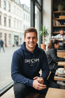 Ein freundlich lächelnder junger Mann in einem dunkelblauen Kapuzenpullover sitzt an einem Tisch direkt am Fenster eines belebten Cafés. Er blickt entspannt in die Kamera, während draußen das städtische Leben vorbeizieht.