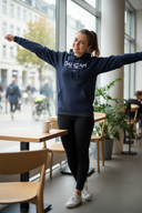 Eine junge Frau in einem dunkelblauen Kapuzenpullover und Leggings steht mit freudig ausgebreiteten Armen in einem modernen Café. Sie lächelt in die Kamera, während neben ihr auf einem Tisch eine dampfende Tasse steht. Durch die großen Fenster ist eine städtische Straße zu sehen.