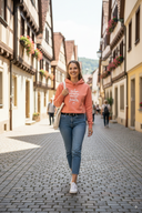 Eine lächelnde junge Frau mit langen braunen Haaren geht auf einer sonnigen Kopfsteinpflastergasse in einer europäischen Altstadt auf die Kamera zu. Sie trägt einen lachsfarbenen Kapuzenpullover, Jeans und eine weiße Stofftasche über der Schulter. Die Gasse ist von historischen Fachwerkhäusern gesäumt.