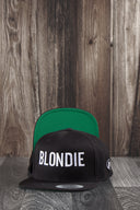 Ein Produktfoto von zwei identischen schwarzen Kappen, die auf einer dunklen Holzoberfläche vor einer rustikalen Holzwand arrangiert sind. Die vordere Kappe zeigt den weißen "BLONDIE"-Schriftzug, während die hintere Kappe aufgestellt ist, um die leuchtend grüne Unterseite des Schirms zu präsentieren.
