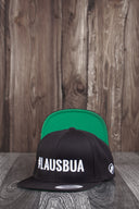 Ein Produktfoto von zwei identischen schwarzen Kappen, die auf einer dunklen Holzoberfläche vor einer rustikalen Holzwand arrangiert sind. Die vordere Kappe zeigt den weißen "#LAUSBUA"-Schriftzug, während die hintere Kappe aufgestellt ist, um die leuchtend grüne Unterseite des Schirms zu präsentieren.