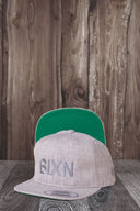 Ein Produktfoto von zwei identischen grauen Kappen, die auf einer dunklen Holzoberfläche vor einer rustikalen Holzwand arrangiert sind. Die vordere Kappe zeigt den Ton-in-Ton gestickten "BIXN"-Schriftzug, während die hintere Kappe aufgestellt ist, um die leuchtend grüne Unterseite des Schirms zu präsentieren.