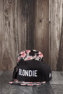 Ein Produktfoto von zwei identischen schwarzen Kappen, die auf einer dunklen Holzoberfläche vor einer rustikalen Holzwand arrangiert sind. Die vordere Kappe zeigt den weißen "BLONDIE"-Schriftzug, während die hintere Kappe die Unterseite des Schirms mit buntem Blumenmuster präsentiert.