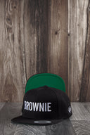 Ein Produktfoto von zwei identischen schwarzen Kappen, die auf einer dunklen Holzoberfläche vor einer rustikalen Holzwand arrangiert sind. Die vordere Kappe zeigt den weißen "BROWNIE"-Schriftzug, während die hintere Kappe aufgestellt ist, um die leuchtend grüne Unterseite des Schirms zu präsentieren.
