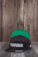 Ein Produktfoto von zwei identischen schwarzen Kappen, die auf einer dunklen Holzoberfläche vor einer rustikalen Holzwand arrangiert sind. Die vordere Kappe zeigt den weißen "#LAUSBUA"-Schriftzug, während die hintere Kappe aufgestellt ist, um die leuchtend grüne Unterseite des Schirms zu präsentieren.